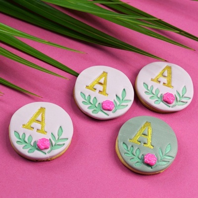 Biscoitos decorados com letra A dourada e flores em fundo rosa
