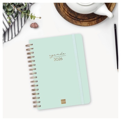 Agenda verde-água com espiral dourado e elástico sobre mesa com jarra branca e toalha riscada