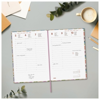 Planner aberto a mostrar calendário semanal sobre mesa com caneca de café, folhas verdes e cadernos.