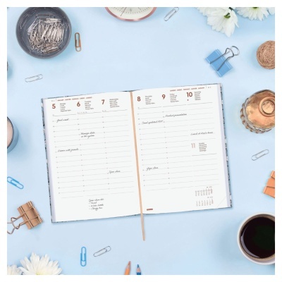 Agenda diária aberta com páginas semanais, marcador, clips e caneca num fundo azul claro