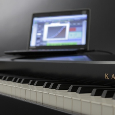 Piano digital preto KAWAI com teclado e computador portátil ao fundo