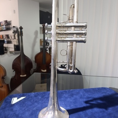 Trompete prateado colocado sobre tecido azul com texto Vam, contrabaixos ao fundo