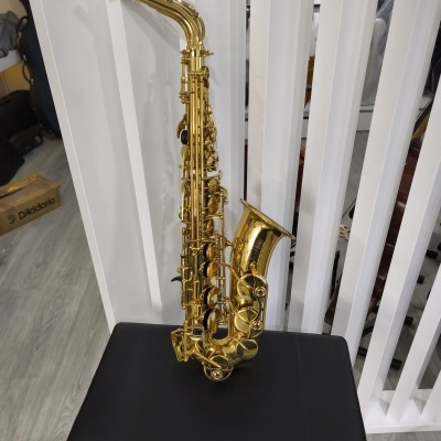 Saxofone alto dourado sobre banco preto com fundo de painéis brancos.