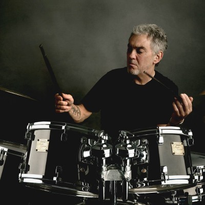 Homem a tocar bateria preta e cromada com baquetas, fundo cinzento