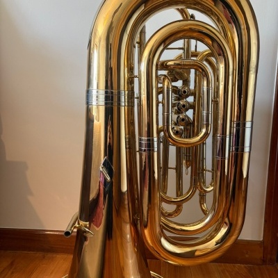 Tuba dourada de metal com válvulas em chão de madeira junto a parede branca