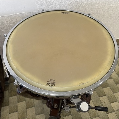 Bumbo de bateria com pele bege e aro metálico prateado em suporte