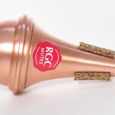 Mute para trompete em metal bronzeado, com logo vermelho e branco, pés em cortiça e uma superfície preta.