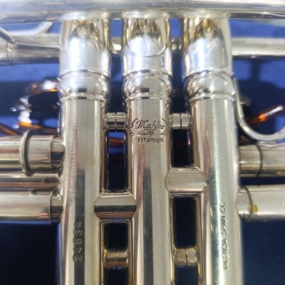 Instrumento musical de metal prateado com três válvulas e inscrições gravadas.