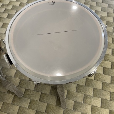 Pele de bateria branca com aro metálico prateado sobre chão de ladrilhos bege e verde