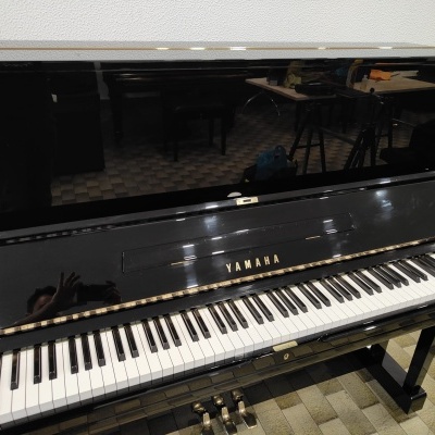 Piano vertical preto brilhante Yamaha com teclado e pedais numa sala com chão em tijoleira.