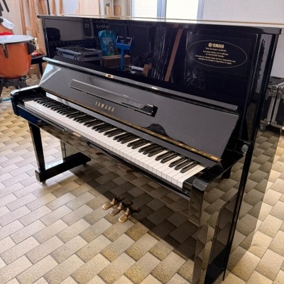 Piano vertical preto Yamaha com teclado e três pedais dourados num chão de azulejos em tons castanhos.