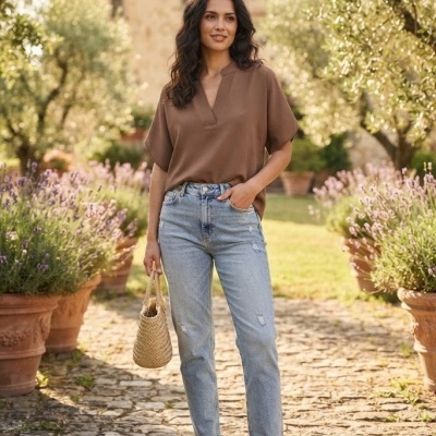 Mulher com blusa castanha, jeans e sandálias castanhas numa alameda com plantas em vasos de barro