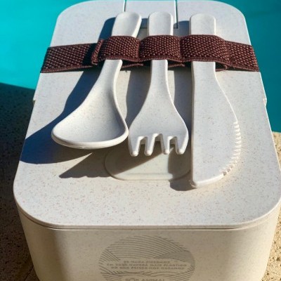 Caixa de almoço bege com talheres de plástico bege presos por correia castanha junto à piscina.