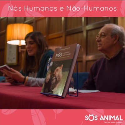 Livro 'Nós Humanos e Não-Humanos' na mesa com duas pessoas sentadas e texto SOS ANIMAL ao fundo