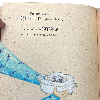 Página de livro infantil com texto e desenho de mãos segurando um saco com um gatinho bebé enrolado