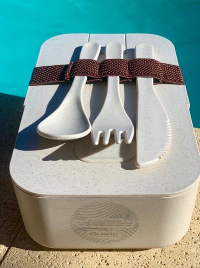 Caixa de almoço bege com talheres de plástico bege presos por correia castanha junto à piscina.