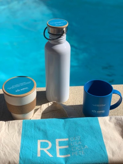 Garrafa metálica branca com tampa de cortiça, caneca azul e copo reutilizável com faixa de cortiça com marca SOS ANIMAL junto a toalha azul perto de piscina