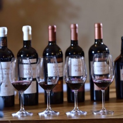 Cinco garrafas de vinho tinto e cinco copos de vinho com vinho tinto parcialmente preenchidos numa bancada de madeira