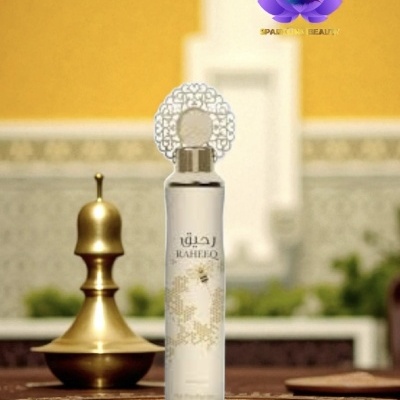 Frasco translúcido branco com texto RAHREEQ e tampa decorativa ao lado de objeto dourado em superfície de madeira