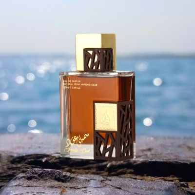 Frasco de perfume com líquido âmbar, tampas e detalhes em madeira castanha, sobre pedra com mar ao fundo