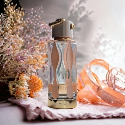 Frasco de perfume em vidro transparente com detalhes bege e dourado numa toalha lilás com flores secas e fita laranja