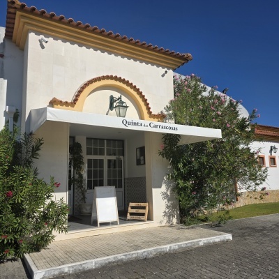 Entrada de prédio com guarda-sol branco e texto Quinta das Carrascosas, plantas floridas ao redor, sob céu azul