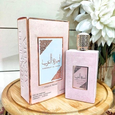Perfume Ambarat Al Arab PRUVE ROSE rosa claro com detalhes dourados e branco sobre base de madeira com flores ao fundo