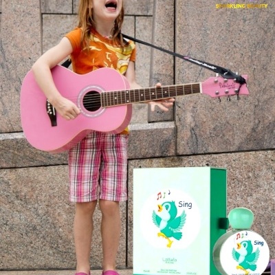 Criança a tocar guitarra cor-de-rosa com frasco e caixa de perfume Sing ao lado.