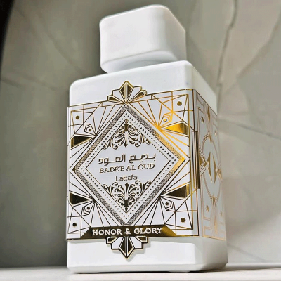 Frasco branco quadrado de perfume com decoração dourada e preta, texto em inglês e árabe.