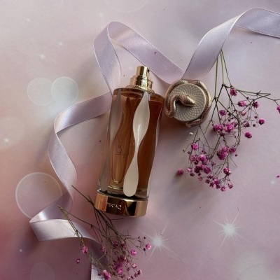 Frasco de perfume âmbar com tampa bege e flores cor-de-rosa em fundo lilás