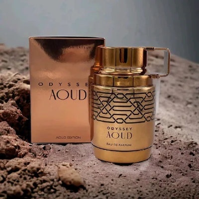 Frasco e caixa dourados de perfume Odyssey Aoud sobre solo arenoso
