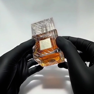 Mãos com luvas pretas seguram garrafa de perfume com líquido âmbar e rótulo em árabe
