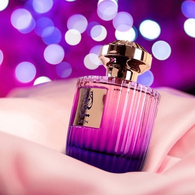 Frasco de perfume rosa e roxo com tampa dourada sobre tecido rosa e fundo de luzes desfocadas roxas.