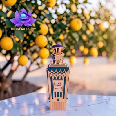 frasco de perfume dourado e azul com texto Elixir SAPPHIRE HAMIDI sobre mármore branco com árvores e frutos amarelos ao fundo