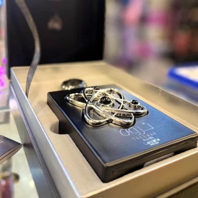 Frasco de perfume preto com texto branco e peça de metal em forma de átomo dentro de caixa branca