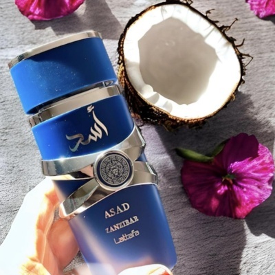 Frasco azul de perfume ASAD ZANZIBAR Latafa, coco partido e flores roxas sobre mesa cinza