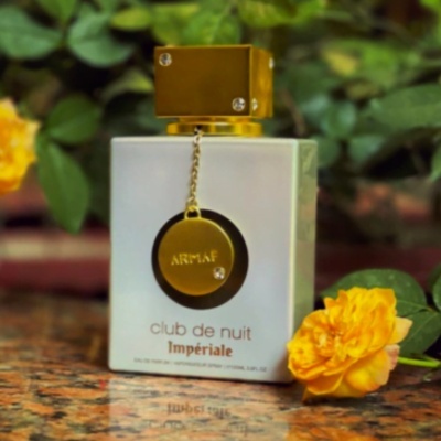 Frasco de perfume branco e dourado com pendente e flores amarelas ao redor