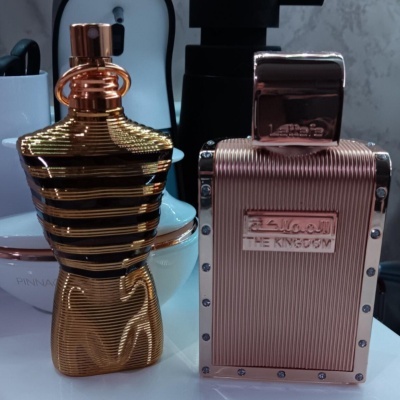 Dois frascos de perfume metálicos, um em forma de torso dourado com listras pretas e outro retangular rosa com texto THE KINGDOM.