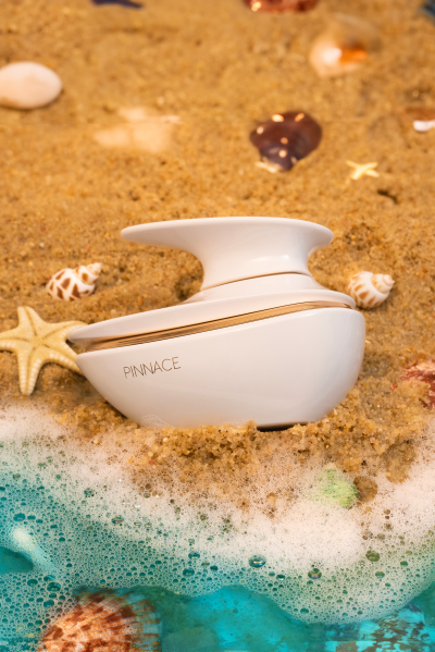 Perfumeiro branco arredondado com tampa, texto PINNACE, na areia com conchas e espuma do mar