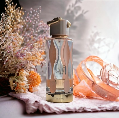 Frasco de perfume em vidro transparente com detalhes bege e dourado numa toalha lilás com flores secas e fita laranja