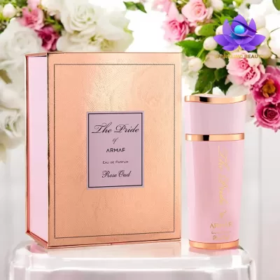 Frasco de perfume rosa claro com detalhes dourados ao lado de caixa rosa e dourada, flores ao fundo