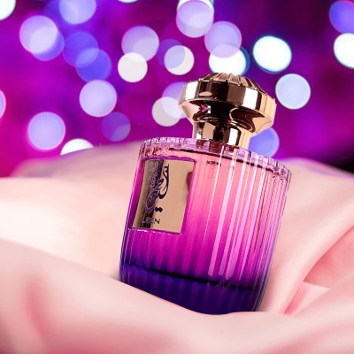 Frasco de perfume rosa e roxo com tampa dourada sobre tecido rosa e fundo de luzes desfocadas roxas.