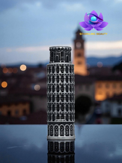 Miniatura metálica da Torre de Pisa sobre superfície reflectora com fundo urbano desfocado e logótipo Sparkling Beauty