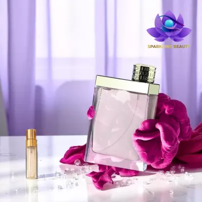 Frasco de perfume transparente com líquido rosa e tampa prateada, pétalas rosa e cristais, cortinas roxas ao fundo.