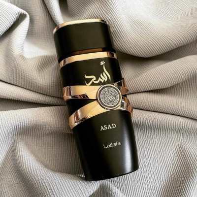 Frasco preto e dourado de perfume ASAD Lattafa sobre tecido cinza