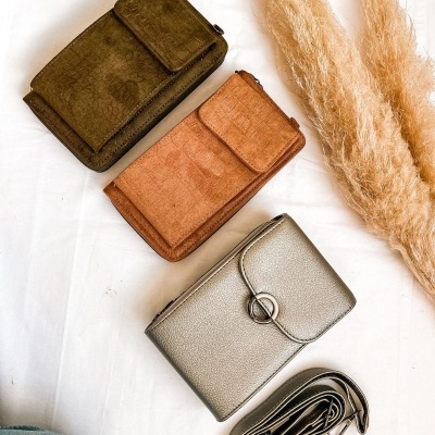 Três carteiras coloridas com textura de pele e uma alça prateada enrolada em fundo branco com plantas secas