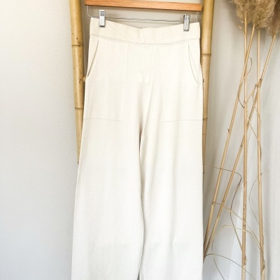 Calças brancas compridas com cintura elástica e bolsos frontais penduradas num cabide de madeira.