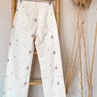 Calças brancas bordadas com flores vermelhas penduradas em cabide de madeira.
