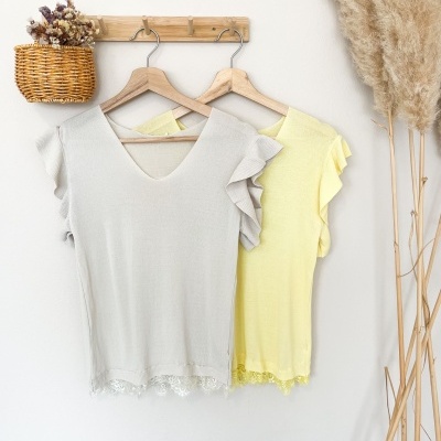 Duas t-shirts femininas bege e amarela com folhos penduradas em cabides de madeira num suporte de parede, com decoração natural ao lado.