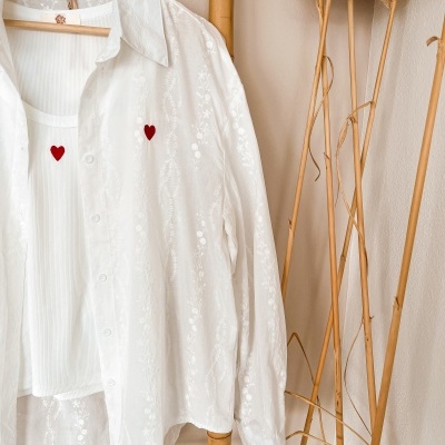 Camisa branca bordada e top branco com corações vermelhos pendurados numa estrutura de madeira junto a hastes decorativas secas